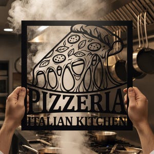 Könnte beinhalten: Ein schwarzes Metallschild mit einem Ausschnittdesign einer Pizzaschnitte mit Peperoni und Blättern sowie dem Text PIZZERIA ITALIAN KITCHEN in einer fetten Serifenschrift. Der quadratische Rahmen wird vor einer Profiküche gehalten.