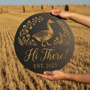 Könnte beinhalten: Ein rundes schwarzes Metallschild mit einer goldfarbenen Gänseillustration, umgeben von Blumen- und Blattmustern. Der Text "Hi There!" ist in Schreibschrift über dem Text "EST. 2023" in einer klaren, serifenlosen Schriftart zu sehen.