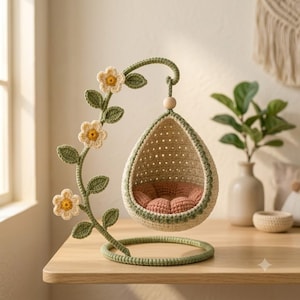 May include: A crochet hanging chair ornament featuring a teardrop-shaped seat with a cream-colored exterior and a dusty rose cushion. The frame is green with a curved vine accent adorned with three cream and yellow crochet flowers and leaves.