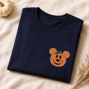 May include: A dark navy blue t-shirt featuring a graphic on the lower right side. The graphic depicts an orange pumpkin shaped like a mouse head with round pumpkin ears, a black smiling face, and a small green leaf on top of the pumpkin stem.