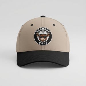 Könnte beinhalten: Eine beige Baseballkappe mit schwarzem Schirm und einem runden Aufnäher auf der Vorderseite. Der Aufnäher zeigt ein gezeichnetes Hochlandrind mit Sonnenbrille und weißer Kappe, umgeben von dem Schriftzug REDMANE GOLF in einem schwarz-weißen Rahmen.