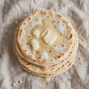May include: A stack of round, flatbreads with golden-brown spots, topped with a melting square of yellow butter and a light dusting of white sugar. The flatbreads are layered on top of each other, showing a soft, cooked texture throughout.