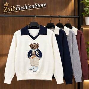 Könnte beinhalten: Ein weißer Langarmpullover mit dunkelblauem Kragen und V-Ausschnitt, der in der Mitte mit einem Teddybären in Jacke und Hose bedruckt ist. Weitere Pullover in Dunkelblau, Grau und Kastanienbraun hängen auf einem Kleiderbügel dahinter.