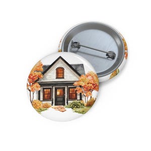May include: A round pin-back button featuring a watercolor illustration of a white house with dark trim, surrounded by autumn trees with orange and yellow foliage. The design includes a stone path leading to the front door and a metal pin back.