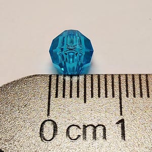 May include: A faceted, translucent bright blue bead positioned against a metal ruler. The bead measures approximately 10 millimetres in diameter, as indicated by the markings on the ruler which show the bead spanning from the zero mark to the ten-millimetre line.