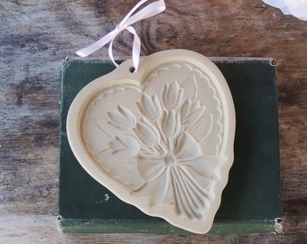 May include: A white ceramic heart-shaped cookie cutter with a raised design of a bouquet of tulips tied with a bow.