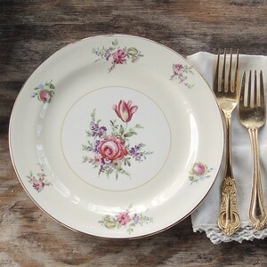 May include: A white ceramic dinner plate with a gold rim and a floral design featuring pink, red, and purple flowers. The plate is set on a wooden table with two gold forks on a white napkin.