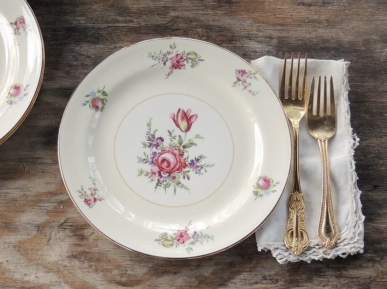May include: A white ceramic dinner plate with a gold rim and a floral design featuring pink, red, and purple flowers. The plate is set on a wooden table with two gold forks on a white napkin.