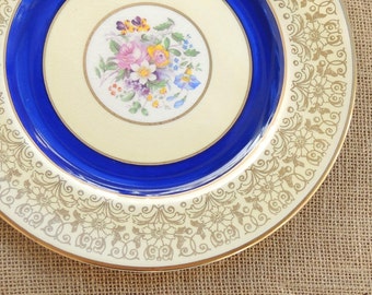 May include: A white ceramic dinner plate with a blue rim and gold floral design. The center of the plate features a floral bouquet in shades of pink, yellow, and blue.