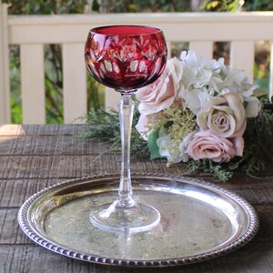 May include: A ruby red crystal wine glass with a decorative cut pattern, set on a silver-colored tray. The glass has a long, slender stem and a round bowl. A bouquet of pink and white flowers is in the background.