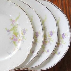 May include: Set of four white ceramic plates with a floral design featuring pink and purple flowers. The plates have a scalloped edge and a gold trim.