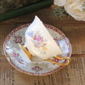 Pode incluir: Uma ch&aacute;vena de ch&aacute; e um pires brancos com uma borda dourada e um padr&atilde;o floral. O pires tem uma borda bord&ocirc; e dourada com um design paisley.