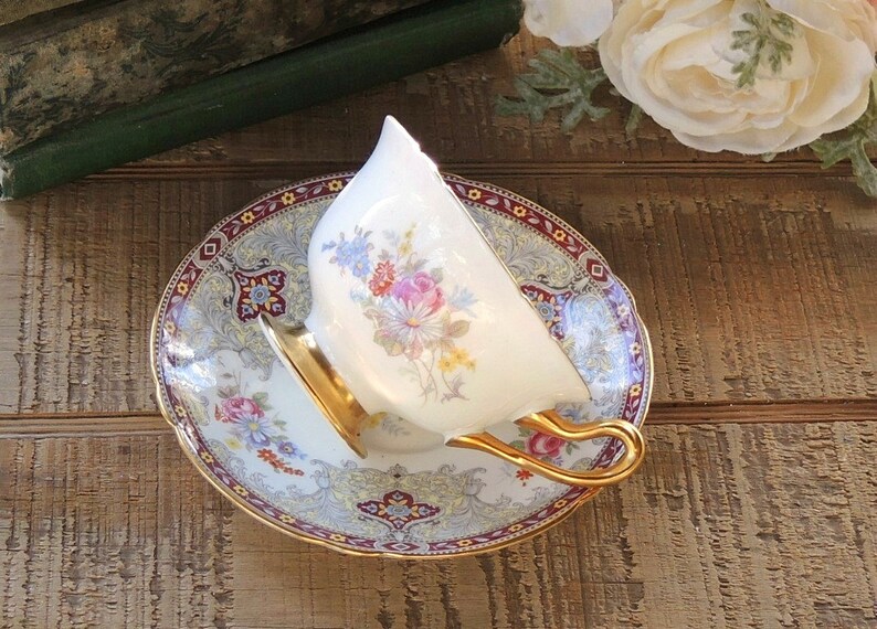 Pode incluir: Uma ch&aacute;vena de ch&aacute; e um pires brancos com uma borda dourada e um padr&atilde;o floral. O pires tem uma borda bord&ocirc; e dourada com um design paisley.