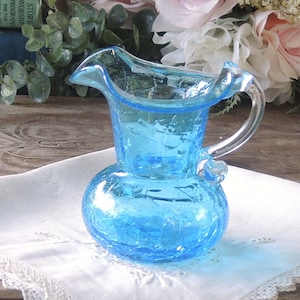 May include: A small, light blue glass pitcher with a textured surface and a clear handle. The pitcher is sitting on a white doily.