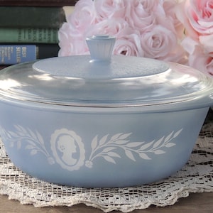 May include: A light blue casserole dish with a white floral design and a clear glass lid. The dish has a white silhouette of a woman's head in the center.