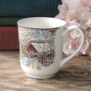 May include: White ceramic mug with a brown and white winter scene featuring a red covered bridge and a snowy mountain. The mug has a brown handle.
