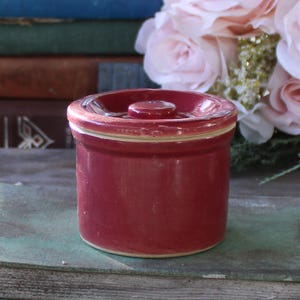 May include: A small, red ceramic crock with a white lid. The crock is sitting on a book.