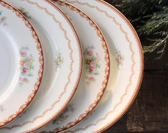 May include: A stack of three white ceramic plates with a floral design. Each plate has a red and gold border with a floral pattern. The plates are arranged on a dark wooden surface.