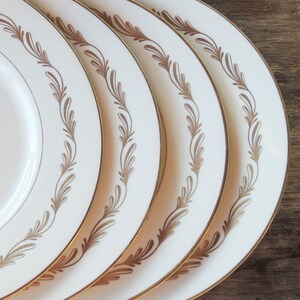 May include: Four white dinner plates with a gold leaf design around the edge. The plates are stacked on top of each other.