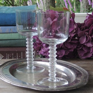 May include: Two clear glass wine glasses with a unique, textured stem design. The glasses are sitting on a silver tray with a decorative border. The glasses are in front of a bouquet of purple flowers.