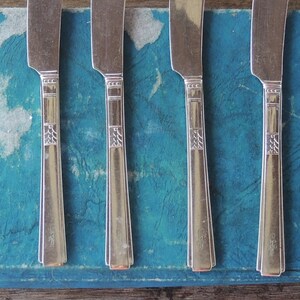 May include: Five silver butter knives with ornate handles are arranged on a blue book cover.