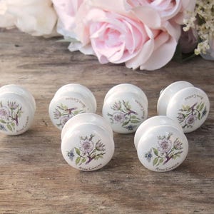May include: Six white ceramic drawer knobs with floral designs and the text "Rosa Canina" printed on the bottom.