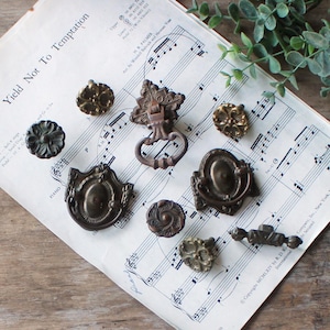 May include: A collection of antique bronze cabinet hardware, including knobs and pulls, arranged on a vintage sheet music page. The hardware features ornate floral and geometric designs, showcasing a rustic aesthetic.