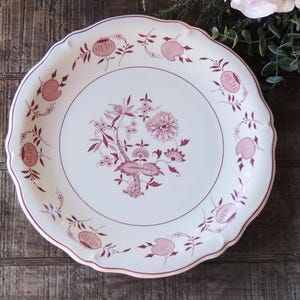 May include: A white ceramic plate with a scalloped edge and a pink floral design. The plate features a central floral motif with smaller floral accents around the rim.