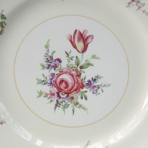 May include: A white ceramic plate with a gold rim features a floral design. The center of the plate has a bouquet of pink, red, and purple flowers with green leaves. The flowers include a single pink tulip.