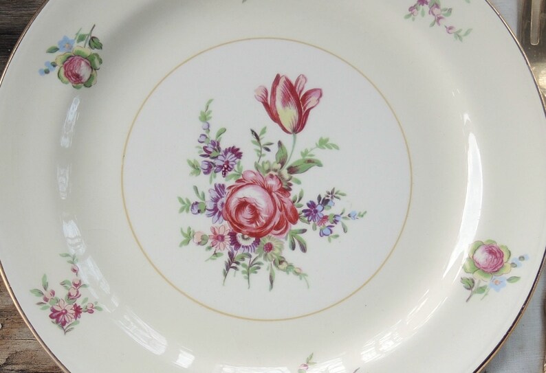 May include: A white ceramic plate with a gold rim features a floral design. The center of the plate has a bouquet of pink, red, and purple flowers with green leaves. The flowers include a single pink tulip.