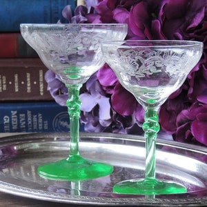 May include: Two vintage green glass champagne coupes with etched floral designs on a silver tray. The coupes are on a wooden surface with a stack of books and purple flowers in the background.