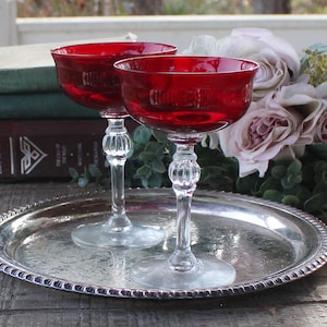 May include: Two ruby red coupe glasses with clear, ornate stems sit on a silver serving tray. The glasses are filled with a dark liquid. A bouquet of flowers and vintage books are in the background.