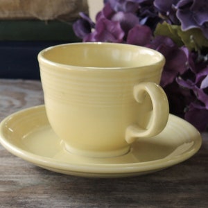 May include: A yellow teacup and saucer set. The cup has a rounded shape and a handle. The saucer is round and has a slightly raised edge.