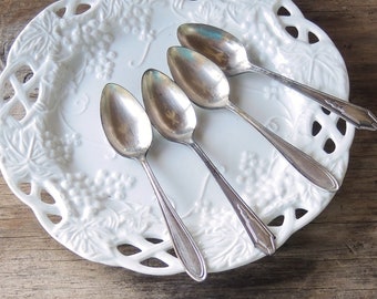 May include: Four silver teaspoons on a white plate with a grapevine design.