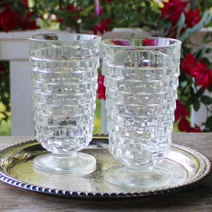 May include: Two clear, textured drinking glasses with a diamond pattern, resting on a silver-colored serving tray. The glasses have a pedestal base. Red roses and a white fence are in the background.