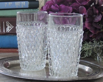 May include: Two clear, diamond-patterned drinking glasses sit on a silver serving tray. The glasses are tall and cylindrical, with a textured surface. The background includes books and purple flowers, suggesting a vintage aesthetic.