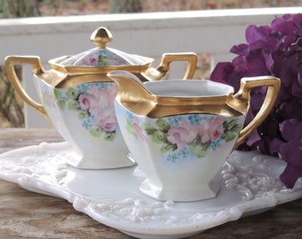 May include: A white porcelain creamer and sugar set with gold trim and floral designs. The set is on a white tray with a floral design. The set is next to a bouquet of purple flowers.