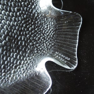 May include: A clear glass fish-shaped dish with a textured surface. The dish has a raised tail fin and the words "Broroo" and "France" are etched on the bottom.