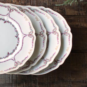 May include: A stack of four white porcelain plates with scalloped edges. Each plate features a delicate floral garland design in shades of purple, green, and gold. The plates are arranged on a dark wooden surface.