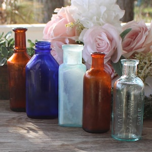 May include: Six vintage glass bottles in various colors, including blue, brown, and clear, with a floral arrangement in the background.