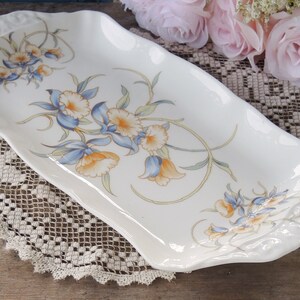 May include: A white ceramic serving tray with a floral design featuring blue, yellow, and green flowers. The tray has a scalloped edge and is perfect for serving snacks or desserts.
