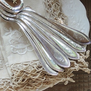 May include: A set of silver-plated spoons with ornate handles, arranged on a beige linen napkin with a lace trim.