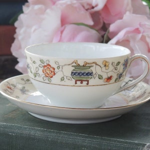 May include: A white teacup and saucer with a floral design featuring a green and blue pot with a gold trim. The teacup and saucer are sitting on a stack of books.