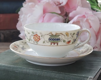 May include: A white teacup and saucer with a floral design featuring a green and blue pot with a gold trim. The teacup and saucer are sitting on a stack of books.
