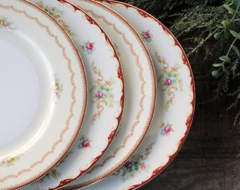 May include: A stack of four white porcelain plates with a floral design. Each plate has a red border and a delicate pattern of pink roses and gold accents. The plates are arranged on a dark wooden surface.