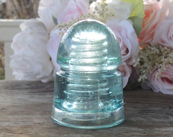 May include: A clear, light blue glass insulator with a rounded top and a cylindrical base. The glass has a textured, ridged design. The background features a bouquet of white and pink flowers, creating a soft contrast to the glass.