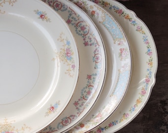 May include: Set of four vintage floral patterned plates in shades of pink, blue, and yellow. The plates are stacked on top of each other, with the largest plate at the bottom and the smallest plate at the top.