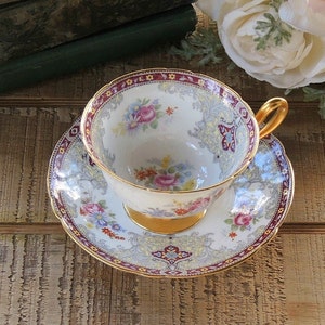 Pode incluir: Uma ch&aacute;vena de ch&aacute; e um pires brancos com um padr&atilde;o floral e guarni&ccedil;&atilde;o dourada. A ch&aacute;vena e o pires t&ecirc;m uma borda festonada e s&atilde;o decorados com uma variedade de flores, incluindo rosas, margaridas e borboletas.
