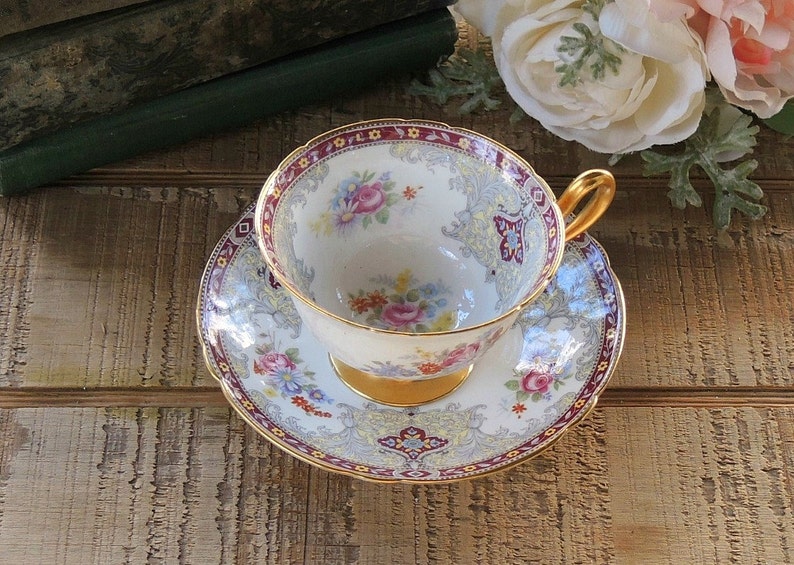 Pode incluir: Uma ch&aacute;vena de ch&aacute; e um pires brancos com um padr&atilde;o floral e guarni&ccedil;&atilde;o dourada. A ch&aacute;vena e o pires t&ecirc;m uma borda festonada e s&atilde;o decorados com uma variedade de flores, incluindo rosas, margaridas e borboletas.