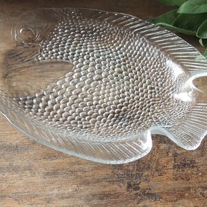 May include: A clear glass fish-shaped serving dish with a textured surface. The dish is designed to resemble a fish with scales and fins.